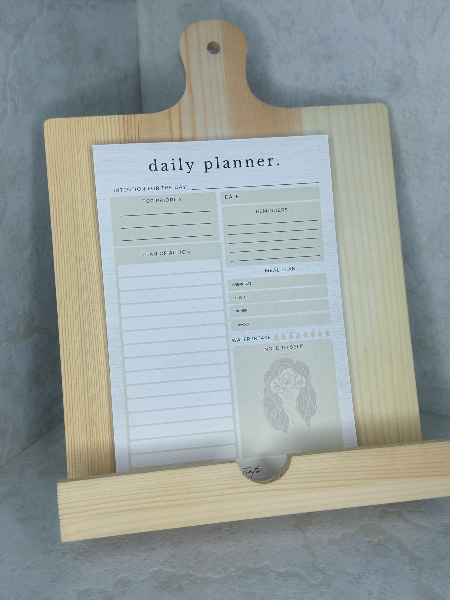 A beige daily planner pad with a layout that includes sections for planning and personal reflections, placed inside a wooden planner stand.