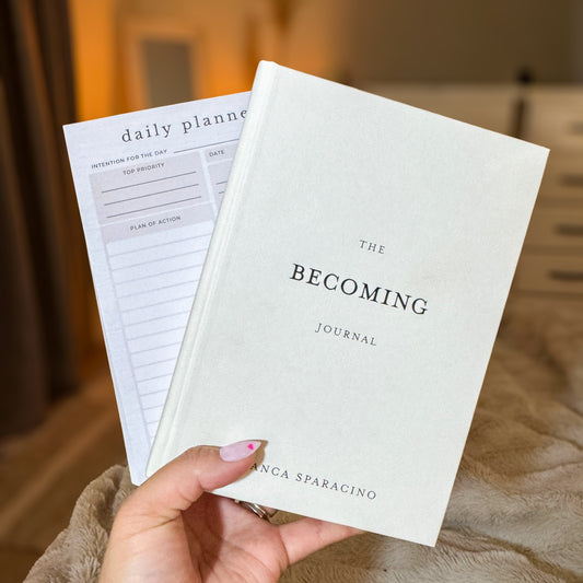 Hand holding 'The Becoming Journal' and Daily Planner Pad with a blurred indoor background